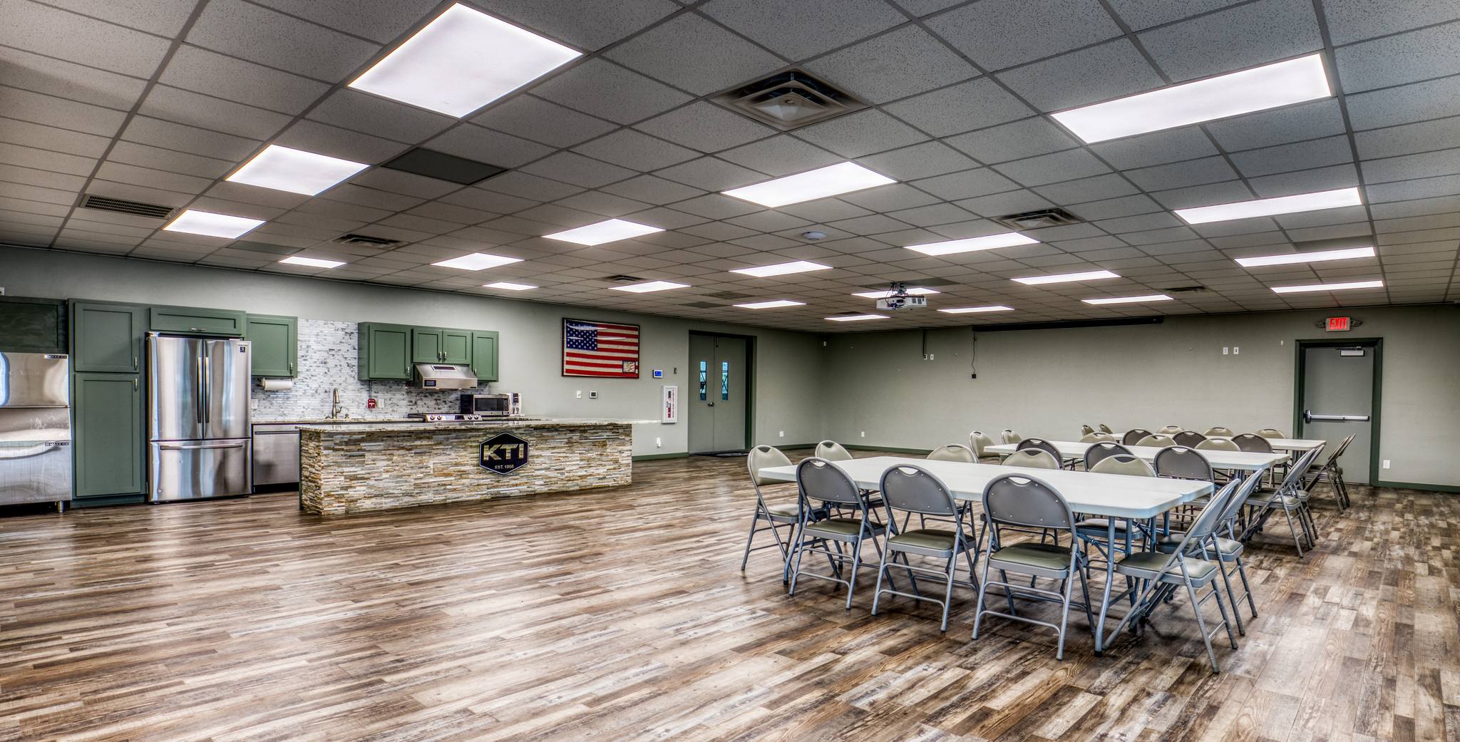 Kenjura Tile Training Center main hall &mdash; wide interior with new wood-look flooring, tables and chairs set up, full kitchen island with KTI hexagon emblem, and meeting-ready layout.